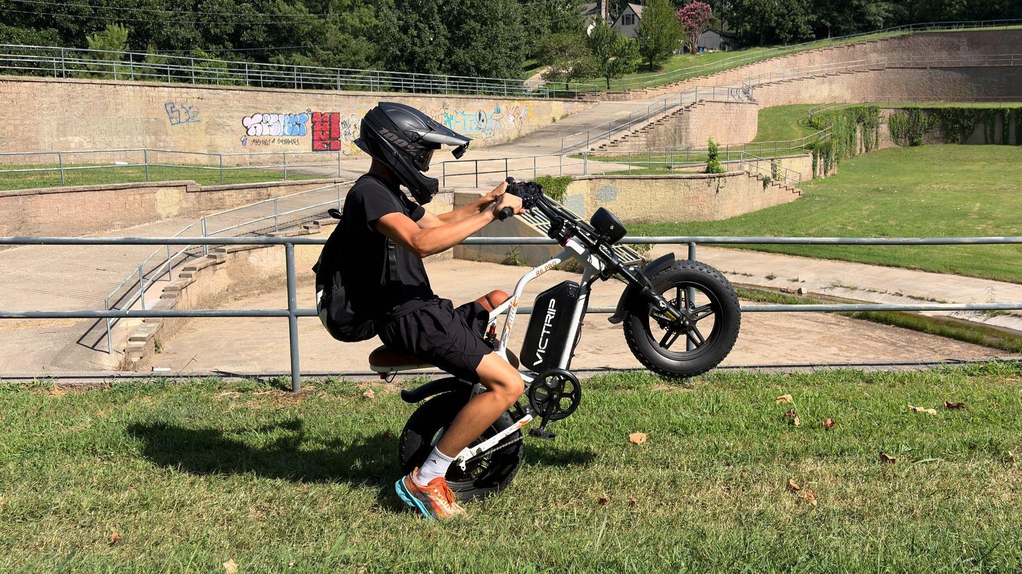 A rider wearing a full-face helmet and backpack pulls a wheelie on a VICTRIP moped-style ebike in a grassy park.