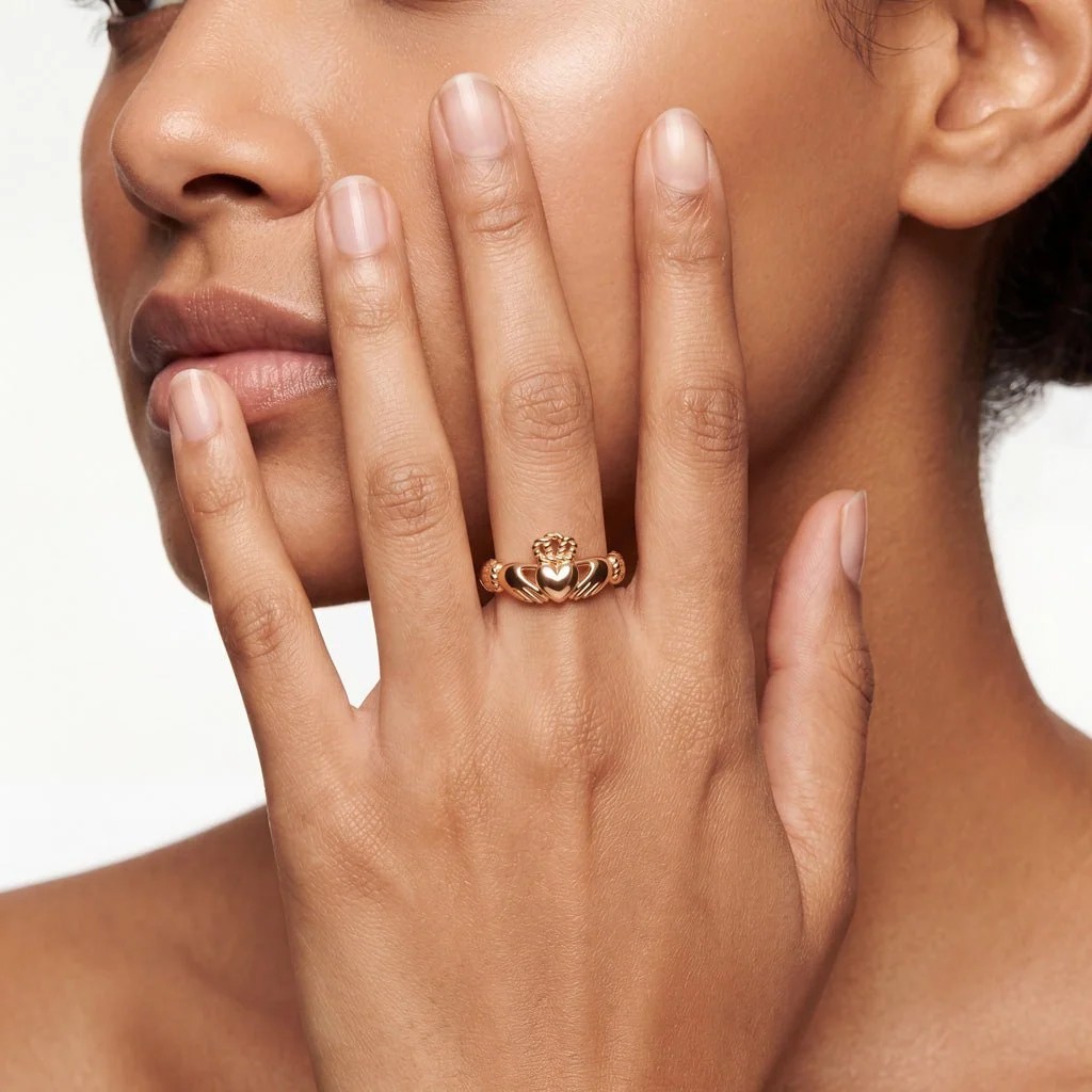 A close-up of a woman's hand showcasing a gold Claddagh ring on her ring finger, near her face.